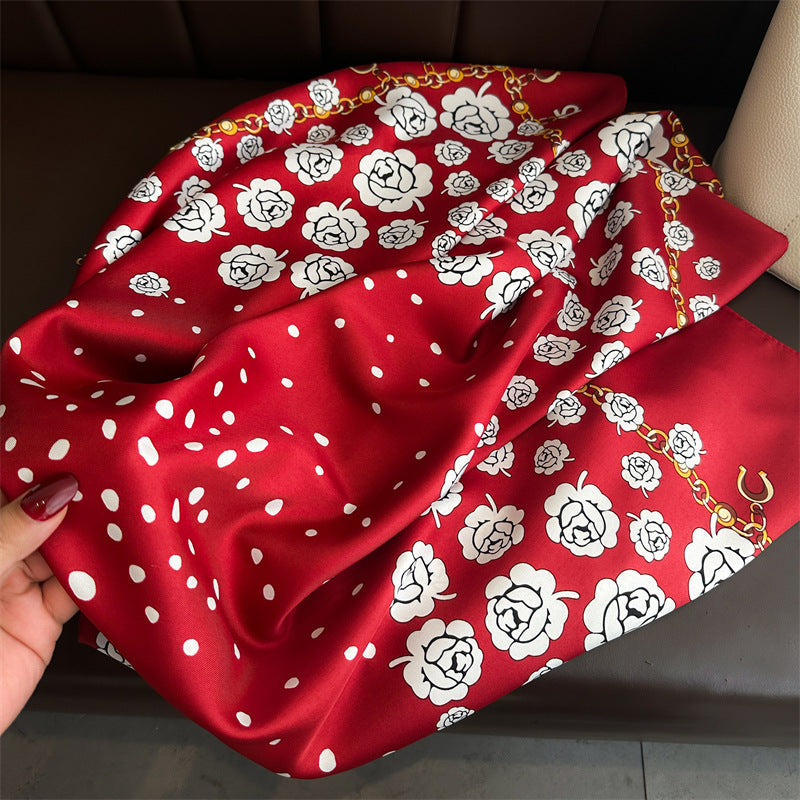 Crimson red silk-feel square scarf held open by model showing satin texture, white camellia rose print, polka dots, and gold chain border