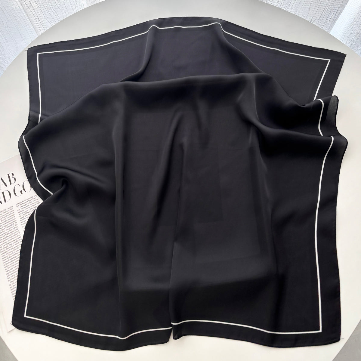 Black silk-feel square scarf with white border trim laid flat and open on white surface - showing full 70cm square size and border detail