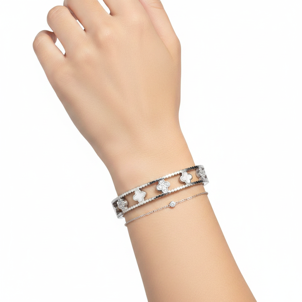 Woman's wrist wearing stainless steel quatrefoil bangle stacked with a delicate chain bracelet, white background